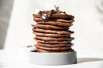 Stack of homemade chocolate pancakes with melted chocolate, ground almond and marshmallow, light background. Direct sunlight and hard shadows.