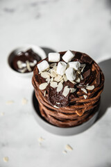 Stack of homemade chocolate pancakes with melted chocolate, ground almond and marshmallow, light background. Direct sunlight and hard shadows.