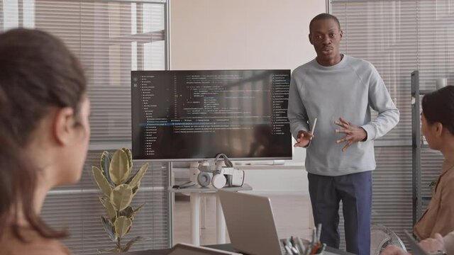 Medium long of excited African IT specialist making presentation, showing computer code on screen to unrecognizable investors sitting in office