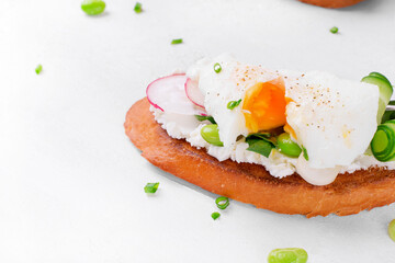 Poached egg on toast with cream cheese, radish, cucumber and edamame beans on the white table. Breakfast sandwich