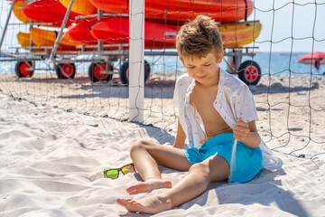 Handsome young boy in blue shorts and white shirt sits on the white sand on the beach. Plays with white sand. High quality photo