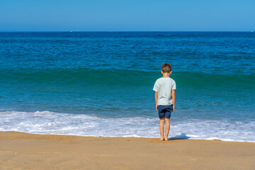 Fototapeta na wymiar One boy stands near the waves on the summer beach. Young teenager, view from the back, looks at the ocean. Summer holiday vacation travel concept. High quality photo
