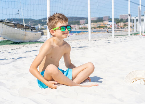 Handsome young boy in blue shorts and sunglasses sits on the white sand on the beach. Plays with white sand. High quality photo