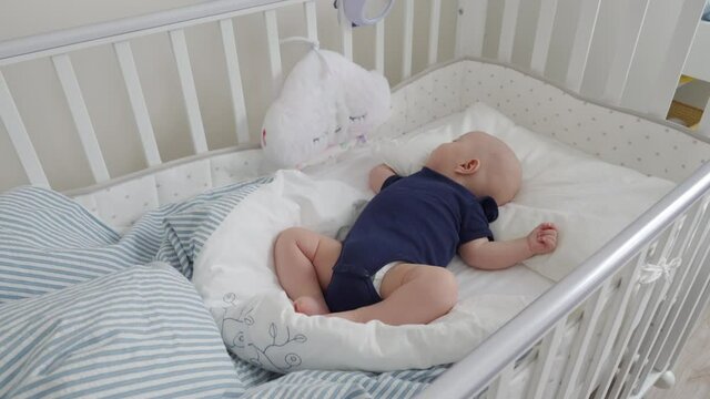 Cute 3 months old baby boy sleeping in a crib under dream cloud lullaby night light, infant in white baby cot closeup. High quality 4k footage