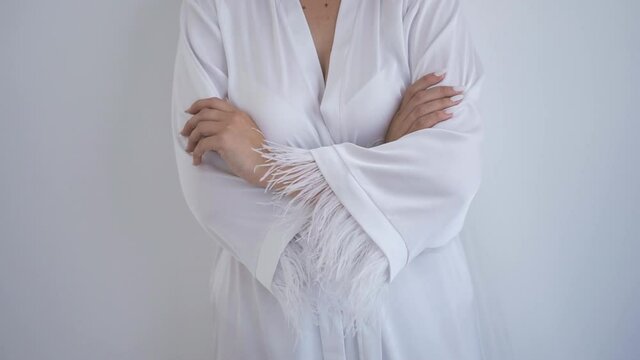 Caucasian bride in a satin robe with feathers on her sleeves stands by the wall and intertwined her arms. Morning of the bride. Photo shoot in a nightgown. Bride's photo shoot