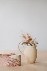 Minimal home decor with dry flowers in a ceramic vase and a cup in female hands. 