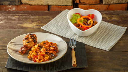 Seashell pasta in a tomato with a grilled chicken leg on a stand and a napkin next to a fork and a vegetable salad.