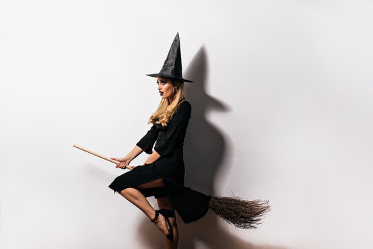 Dreamy young witch flying on broom in halloween. Indoor shot of debonair blonde wizard posing on white background.