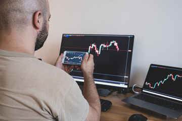 The young man is studying the market charts on both the phone and the computer screen.