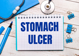 On a light wooden background, a stethoscope, a blue notepad, blue paper clips, a blue marker and a sheet of paper with the text STOMACH ULCER. Medical concept