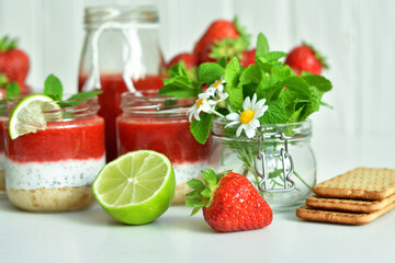 Homemade multi-layer dessert with strawberry and cream cheese in a glass jar. Ripe fresh green lime and mint on white background.