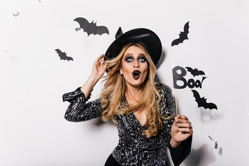 Dreamy caucasian girl in wizard hat expressing surprised emotions. Indoor photo of happy european woman in witch attire celebrating halloween.