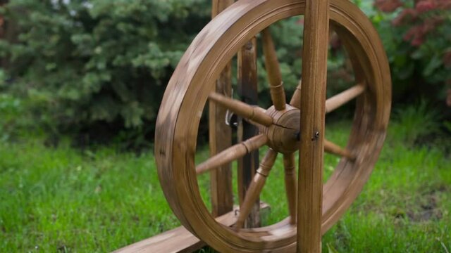 Wooden spinning wheel is on grass in yard. Tool on which threads were spun. Subject of folk life for spinning. Spinning wheel for yarn thread. Ancient craft. Hand touches wheel and it starts spinning