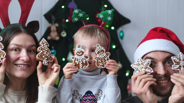 Christmas family portrait. Cheerful mother, father and son having fun near alternative christmas tree holding gingerbread cookies near eyes.