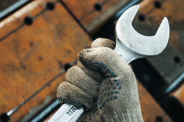 A worker's hand in a glove holds a wrench in a clenched fist, against a background of iron, rusty objects. 