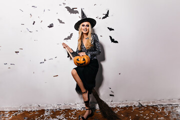 Good-looking woman in hat posing with bats on background. Cheerful blonde lady with halloween pumpkin having fun at vampire party.