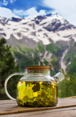 Hot tea with herbs in glass teapot on table in front of high mountains with snow peaks and blue sky with clouds outdoors.