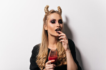 Adorable vampire girl posing in halloween. Indoor photo of sensual lady with black makeup drinking blood.