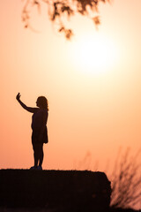 Fototapeta premium Profile silhouette of a woman in a skirt taking a selfie with a smartphone on top of a bale of straw with the sunset behind her. Vertical image.