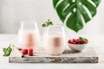 Two glasses with a Lassi drink with raspberries, mint and a bamboo tube on a white wooden board. Light background with a monstera leaf.