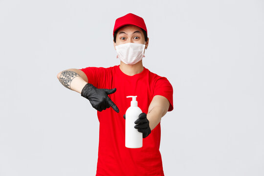 Excited Delivery Guy Pointing At Hand Sanitizer Bottle And Looking Camera, Ensure Clients And Employees Safety During Covid-19 Virus Outbreak. Courier In Medical Mask And Gloves Working