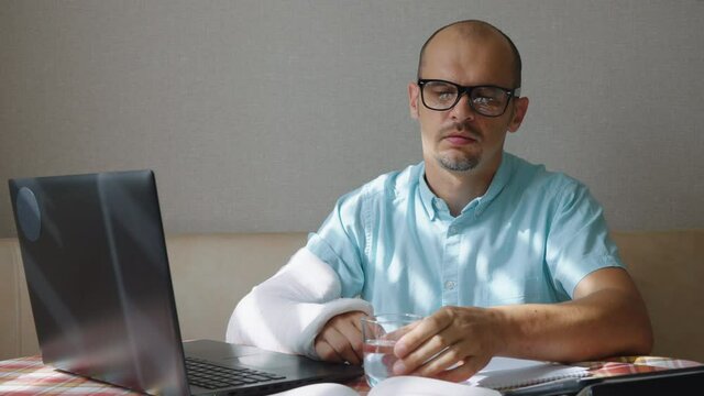 Bearded man in eyeglasses with arm in plaster cast sitting in front of laptop and taking pills. Businessman taking pain killer medicine after accident. Concept of healthcare