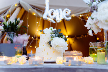 Two tiered wedding cake decorated with fresh peony flowers and edible gold on the wooden table with flowers, candles. Outdoor wedding with lights background. Wedding reception, wedding inspiration