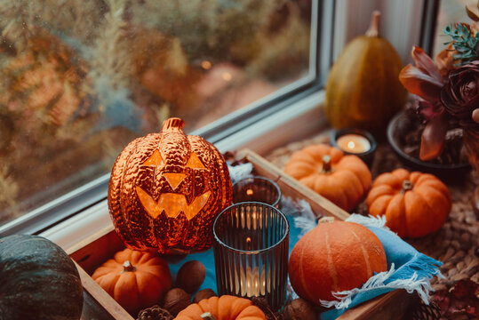 Halloween cozy mood composition on the windowsill. Lighting jack-o-lantern, decorative pumpkins, cones, candles on wooden tray and straw napkin, warm plaid. Hygge halloween home decor. Selective focus