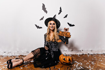 Long-haired vampire girl sitting on the floor with pumpkin. Inspired lady in witch hat posing with...