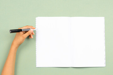 Kid hand holding pen in left hand to write on blank open notebook over pastel green background....