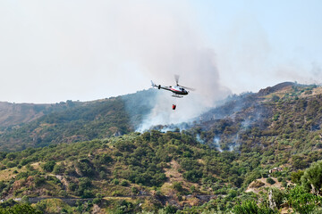 Fototapeta na wymiar Fire helicopter dropping water from a bucket on a forest fire in the mountains. Forest fire is an environmental disaster. Deforestation.