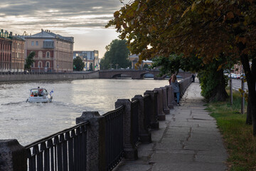 The end of the day or evening on the embankment of the city canal. Stone embankment in the shade of trees, where people rest. Speedboat goes along the river with tourists and passes by city buildings