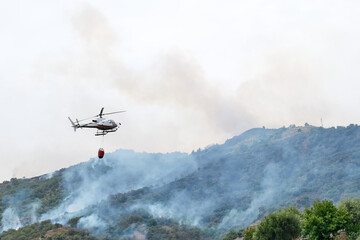 Obraz na płótnie Canvas Fire helicopter dropping water from a bucket on a forest fire in the mountains. Forest fire is an environmental disaster. Deforestation.