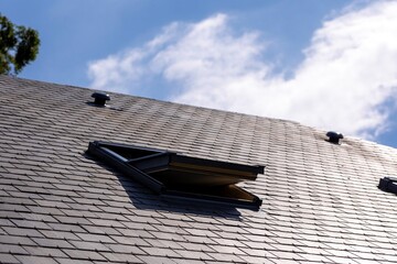 A portrait of an opened skylight window in a slate roof on a sunny day with a blue sky with white...