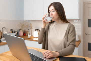 Pretty caucasian girl in beige t-shirt and grey jacket took a break from work and drinks water. At the table laptop and notepad. Home office concept. Woman shopping online or surfing in web.