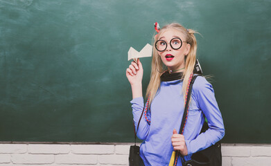 Portrait of a teen female student. Cute teen girl student. Attractive young girl holding backpack and paper plane, standing against blackboard.