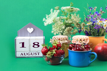 Fototapeta na wymiar Calendar for August 18 : the name of the month of August in English, cubes with the number 18, bouquets of wild flowers, jars of jam, strawberries and cherries in cups, green background
