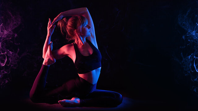 Yoga workout in neon light. Slim young woman yogi does yoga asana Eka pada raja kapotasana. Studio, black background, red and blue neon light. Smoke from an incense stick.