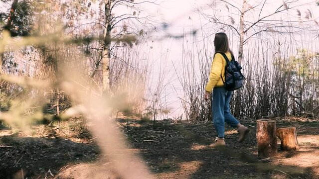 Tourism, nature walks. Young woman with a large backpack and a yellow jacket walks up to the river, stops and enjoys the beautiful view and fresh air. Slow motion, 4k