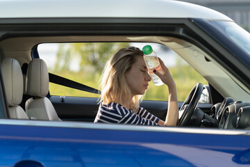 Exhausted woman driver feeling headache, sitting inside her car, applies bottle of water to forehead, hot weather. Tired female stop after driving car in traffic jam. Blood pressure, heat concept. 