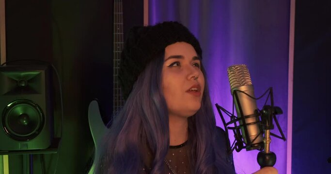 Rock n roll punk girl with tattoo and black beanie singing, recording song with atmospheric vocal in a record label booth. Close up shot in slow motion. Color hair female and night dress with guitar