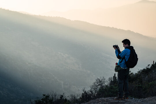 Young man dressed in a blue windbreaker taking pictures with his camera at sunset with the mountains in the background