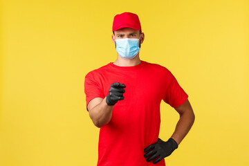 Fototapeta na wymiar Express delivery during pandemic, covid-19, safe shipping, online shopping concept. Confident courier in red uniform, medical mask and gloves, pointing finger camera, promote logistic company