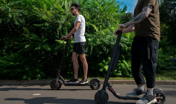 Two Friends Driving Electric Scooter In Park And Having Fun At Sunny Day.