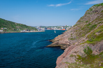 A footpath, hiking trail, or path along a hillside. The cliff is rocky with grass patches. The city of St. John's, Newfoundland, is in the background on a sunny day. The sky is bright blue. 