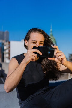 young brazilian man with long curly hair taking a photo with his smart phone, while smiling. vertical photo