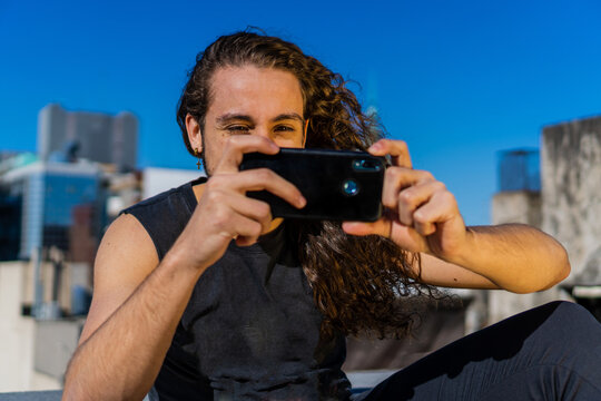 young Brazilian man with long hair and blond curls taking a photo with his smart phone, smiling.