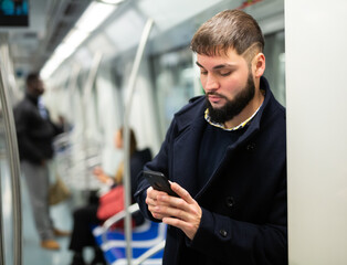 Young focused man browsing and typing messages on phone on way to work in modern metro car