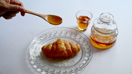 Closeup of Male hand pouring honey from a wooden spoon onto the croissant in a glass dish with honey in a glass jar and a small glass on white background. Healthy food concept, selective soft focus.