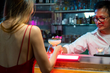 Asian woman sitting at bar counter drinking cocktail and talking to barman in nightclub. Male mixologist bartender preparing alcoholic drink to customer. Small business bar and city nightlife concept
